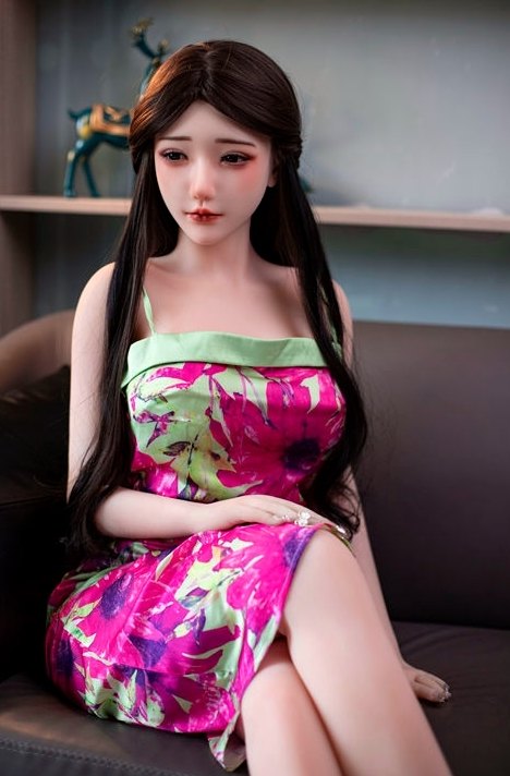 Aadhira 158cm Japanese TPE sex doll seated on a leather sofa in a floral slip dress