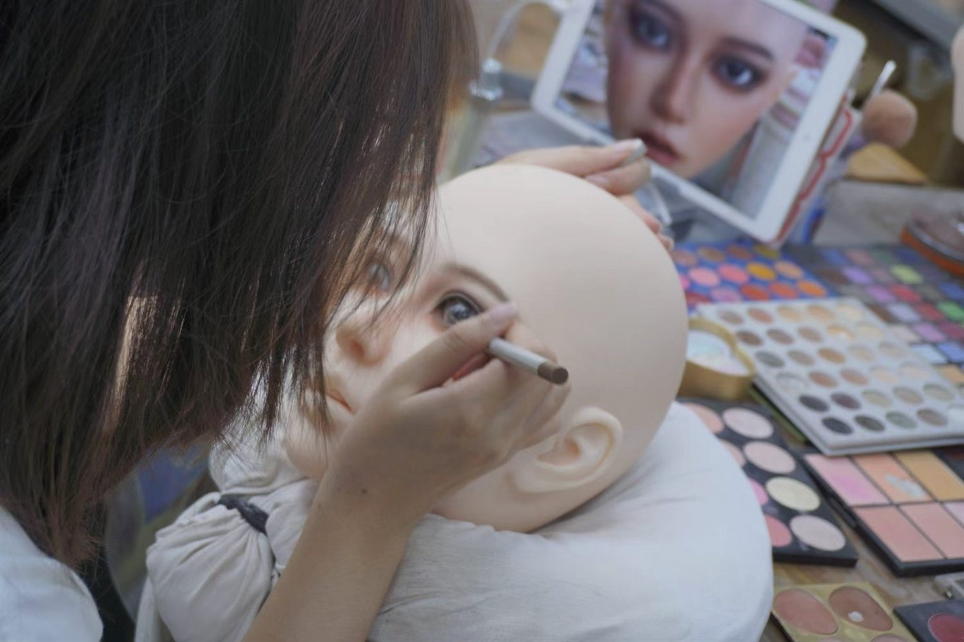 Artist hand-painting brows on a silicone sex doll head against a reference tablet